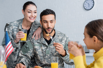 Canvas Print - Smiling parents in military uniform looking at child near american flag during breakfast at home