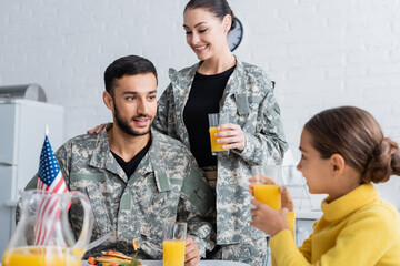 Poster - Smiling parents in military uniform looking at daughter near american flag during breakfast at home