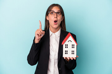 Wall Mural - Young business woman holding a toy home isolated on blue background pointing upside with opened mouth.