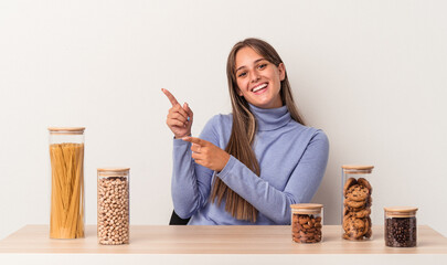 Wall Mural - Young caucasian woman sitting at a table with food pot isolated on white background excited pointing with forefingers away.