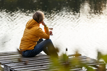 Wall Mural - Woman in a yellow jacket relaxing on a wooden pier on a lake with a glass of rose wine watching fall sunset alone.