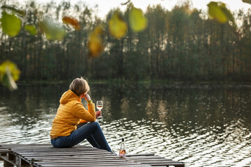 Wall Mural - Woman in a yellow jacket relaxing on a wooden pier on a lake with a glass of rose wine watching fall sunset alone.