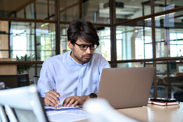 Young Indian businessman using computer working online, doing research, taking notes, watching business training webinar in office, having virtual chat conference meeting looking at laptop at work.