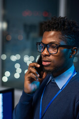 Young confident businessman in smart casualwear and eyeglasses consulting client by mobile phone in office environment