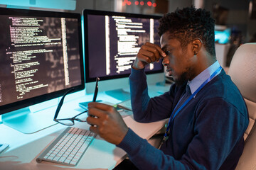 Poster - Young tired African businessman sitting by workplace late at night while trying to concentrate and continue working