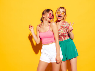 Two young beautiful smiling blond hipster female in trendy summer clothes. Sexy carefree women posing near yellow wall in studio. Positive models having fun. Cheerful and happy. In sunglasses