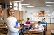 © littlewolf1989 - Portrait of school teacher with notebook standing in classroom and children sitting in background.