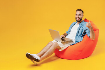 Wall Mural - Full body young smiling fun cool happy man wear blue shirt white t-shirt sit in bag chair hold use work on laptop pc computer show thumb up gesture isolated on plain yellow background studio portrait