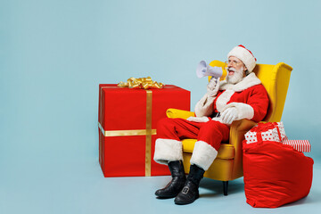 Full body old Santa Claus man in Christmas hat red suit clothes sit in chair scream in megaphone isolated on plain blue background studio. Happy New Year celebration merry ho x-mas holiday concept