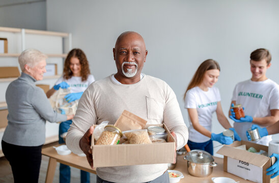 charity center. happy black senior man holding box with donations food, looking and smiling at camer
