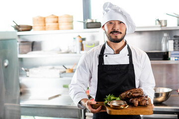 Smiling asian chef holding cutting board with roasted meat and greens in kitchen