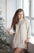 © Dina - Beautiful young caucasian brunette woman standing near window and decorated christmas tree at home on holiday season