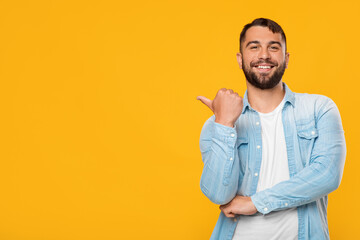 Smiling adult attractive caucasian male points finger towards empty space, isolated on yellow background