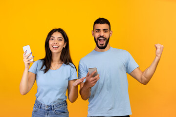Online lottery. Overjoyed young arab man and woman rejoicing success with their smartphones, yellow background