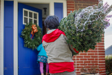 A Man Carries Christmas Tree Into Home With Daughter In Background