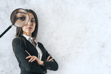 Poster - Portrait of attractive young european businesswoman with magnifier on eyes standing on white concrete wall background with mock up place. Search and discovery concept.