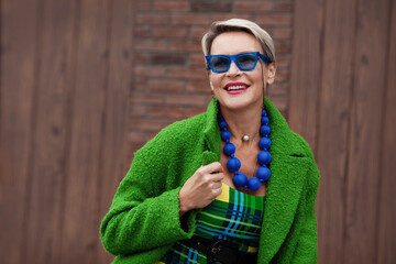 Portrait of beautiful adult woman with stylish short hair having beautiful smile and wearing bright green coat, fashion accessories, blue sunglasses and beads