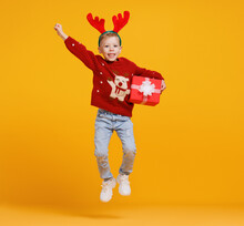 Excited Little Boy In Christmas Sweater And Deer Antlers Jumping Up In Air With Xmas Gift Box