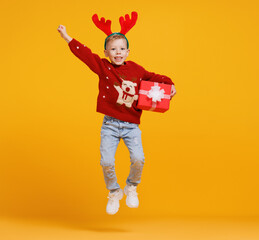 Excited little boy in Christmas sweater and deer antlers jumping up in air with Xmas gift box