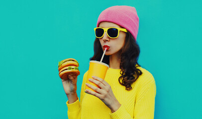 Portrait of stylish young woman drinking juice with burger fast food on blue colorful background