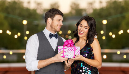 Poster - valentines day, birthday, anniversary and holidays concept - happy couple with gift box at rooftop party over lights on background