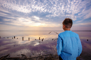 Rear view of a cute teenage boy in summer clothes standing on an amazing pink lake with salt and healing mud. Sunset sky.