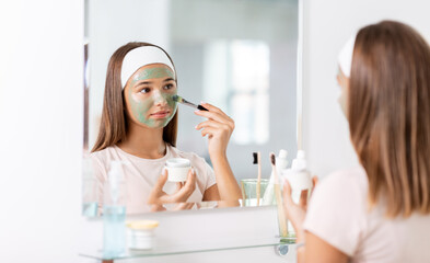 Sticker - beauty, skin care and people concept - teenage girl with brush applying clay mask to face and looking in mirror at bathroom