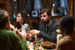 © AnnaStills - Young bearded man in suit talking to people while sitting at dining table during dinner