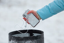 Closeup of young female frozen hand throwing her broken smartphone and earphones away into a trash during a walk in the snowy winter park
