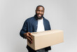 © Vadim Pastuh - Glad young African-American man holding cardboard box, smiling and looking at camera isolated on gray background, delivery boy. Happy black guy moved in new house, relocation concept