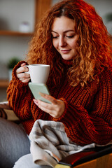 A cute ginger girl with curly hair is sitting in the chair at home in the morning, texting messages on the phone and drinking her coffee.