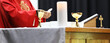 A Catholic Priest about to break the bread host while celebrating blessed Holy Communion at Mass. Wearing a red gown vestment and surrounded by chalice cross crucifix and bible