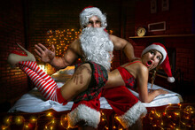 Portrait Of Santa Claus Sitting In The Bed With Sexy Girl In Santa Hats And Slaping Her