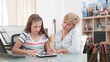 © DC Studio - Mother sitting beside daughter during homeschooling helping her with literature lesson, reading together online book sitting at desk. Child studying using elearning school platform