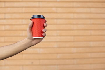 Drinks, lifestyle and people concept - Close-up of a red cup of coffee with a black lid held by a African-American male hand on a brick wall background with copy space