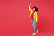 © ViDi Studio - Full size body length bright young curly black latin woman 20s years old wears yellow jacket go move stroll meet greet waving hand as notices someone isolated on plain red background studio portrait.