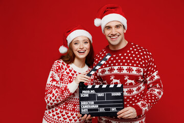 Young smiling couple friends two man woman 20s in sweater hat holding classic black film making clapperboard isolated on plain red background studio portrait. Happy New Year 2022 celebration concept.