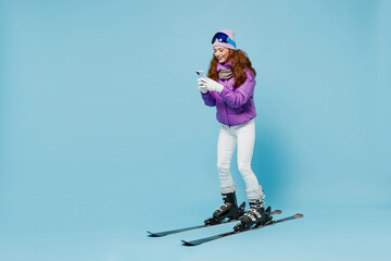 Wall Mural - Full size skier woman in warm purple padded windbreaker jacket ski goggles mask spend extreme weekend in mountains use hold mobile cell phone browsing internet isolated on plain blue background studio