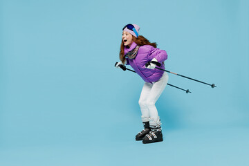 Wall Mural - Full body side view skier fun cool caucasian woman wear warm purple padded windbreaker jacket goggles mask spend extreme weekend in mountains skiing isolated on plain blue background studio portrait.