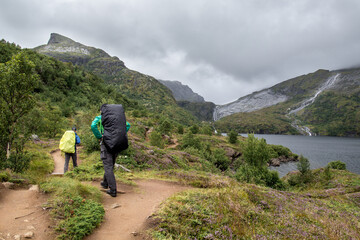 two hiker with backpacks on a path near a lake surrounded by high mountains and cloudy rainy sky on 