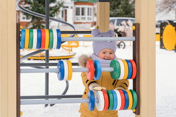 A baby in a yellow jacket aged 12-17 months plays with colorful wooden bills on the playground. Wintertime Russia