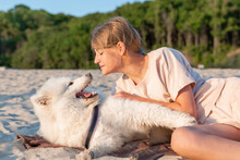 A Teenage Girl Gently Looks At Her Dog Husky Samoyed, They Play Lying On The Sandy Beach.  Friendship With A Pet, Children And Animals Concept.
