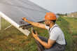 Competent technician in safety helmet and glasses measuring amperage of solar panels with multimeter. Indian man repairing photovoltaic cells outdoors. Green energy concept.