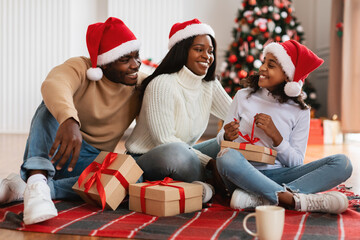 Wall Mural - Cheerful black girl celebrating Christmas with family unwrapping gift box