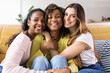 © Xavier Lorenzo - Three united beautiful smiling women sitting together on couch - Portrait of young multiracial girl friends hugging each other and relaxing at home - Friendship, youth and millennial people concept