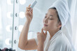 Beautiful girl in bathrobe and turban towel on her head, spray mineral water on face in front of the mirror, women's lifestyle