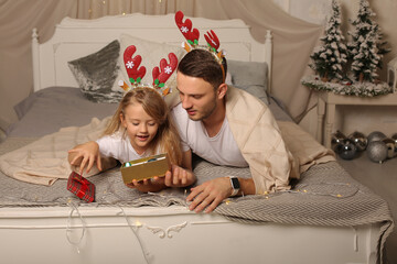 Wall Mural - a father is lying on a bed with a little daughter in a deer antler rim watching a Christmas gift box