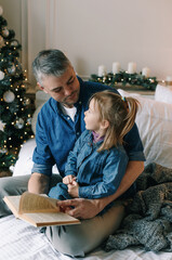 Wall Mural - An attentive father reads a Christmas tale for a happy girl sitting on a bed near a decorated Christmas tree