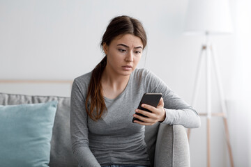 Canvas Print - Sad young female sits on sofa look at smartphone and reading message at home, frustrated by received notification