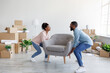 Cheerful young african american female and male carry armchair to new apartment with cardboard boxes